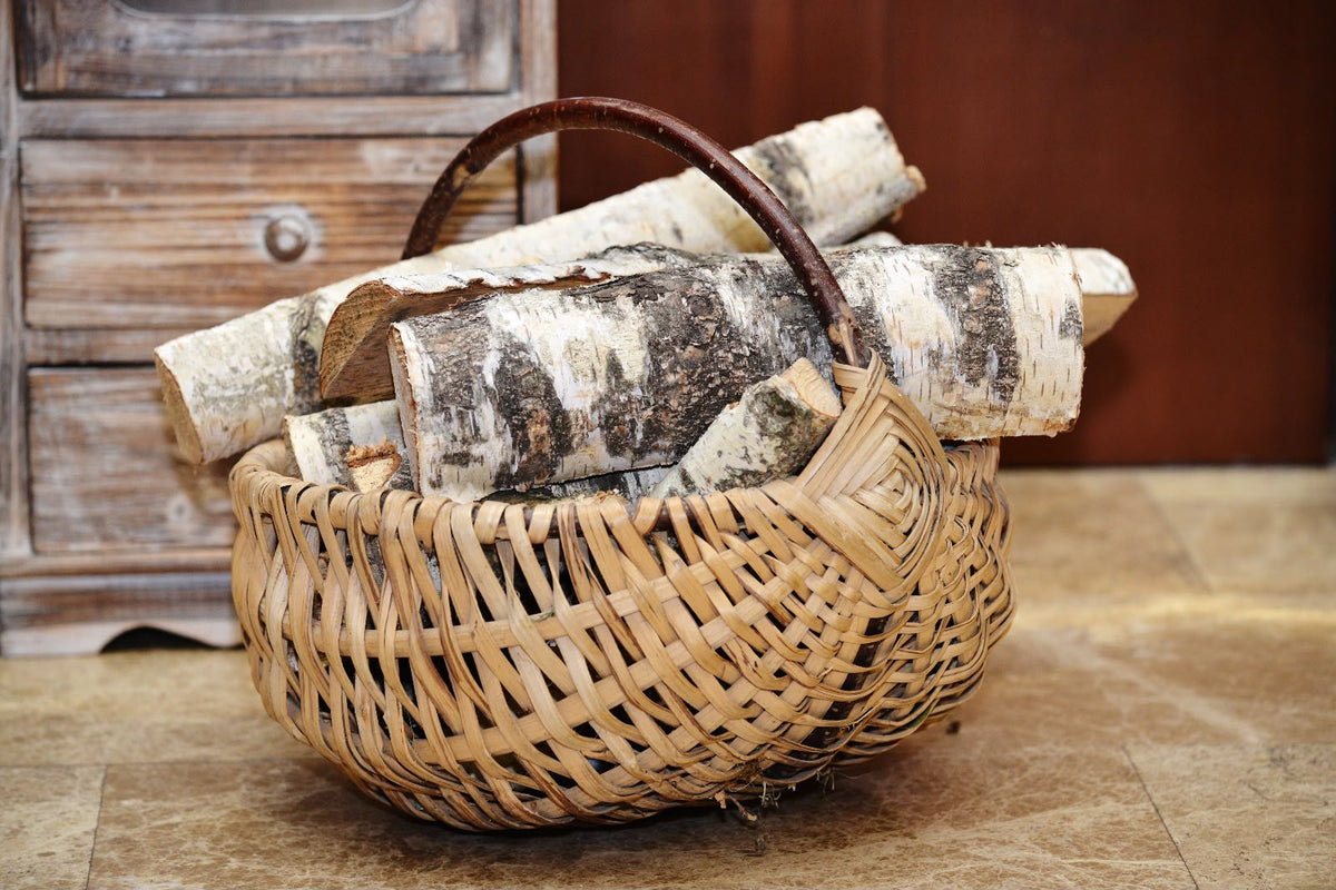 A woven natural-fiber basket with a dark brown handle, filled with cut birch logs, set on a stone floor in front of a rustic wooden cabinet.