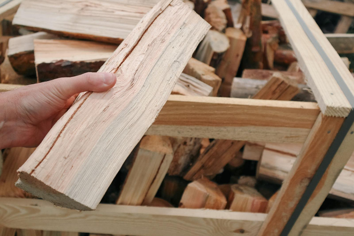 A hand holding a Birch firewood log in front of a wooden crate filled with stacked logs .
