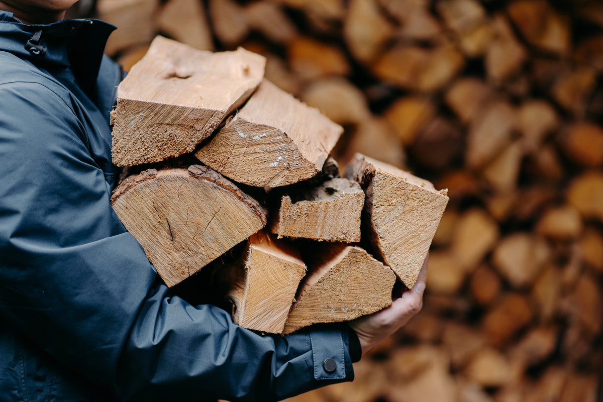 A man carrying a bundle of oak firewood nets with a firewood log store in the background.
