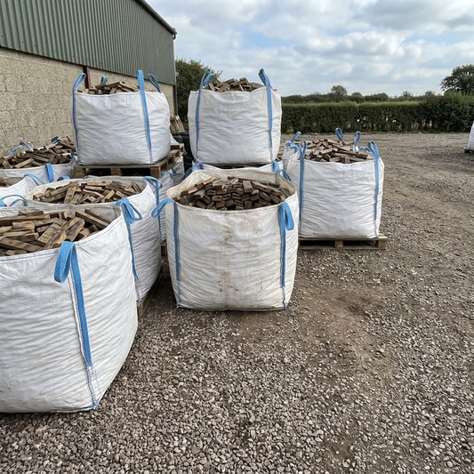 Several large white bulk bags filled with whisky barrel stave firewood stacked on pallets, photographed outdoors on a gravel surface beside a building.